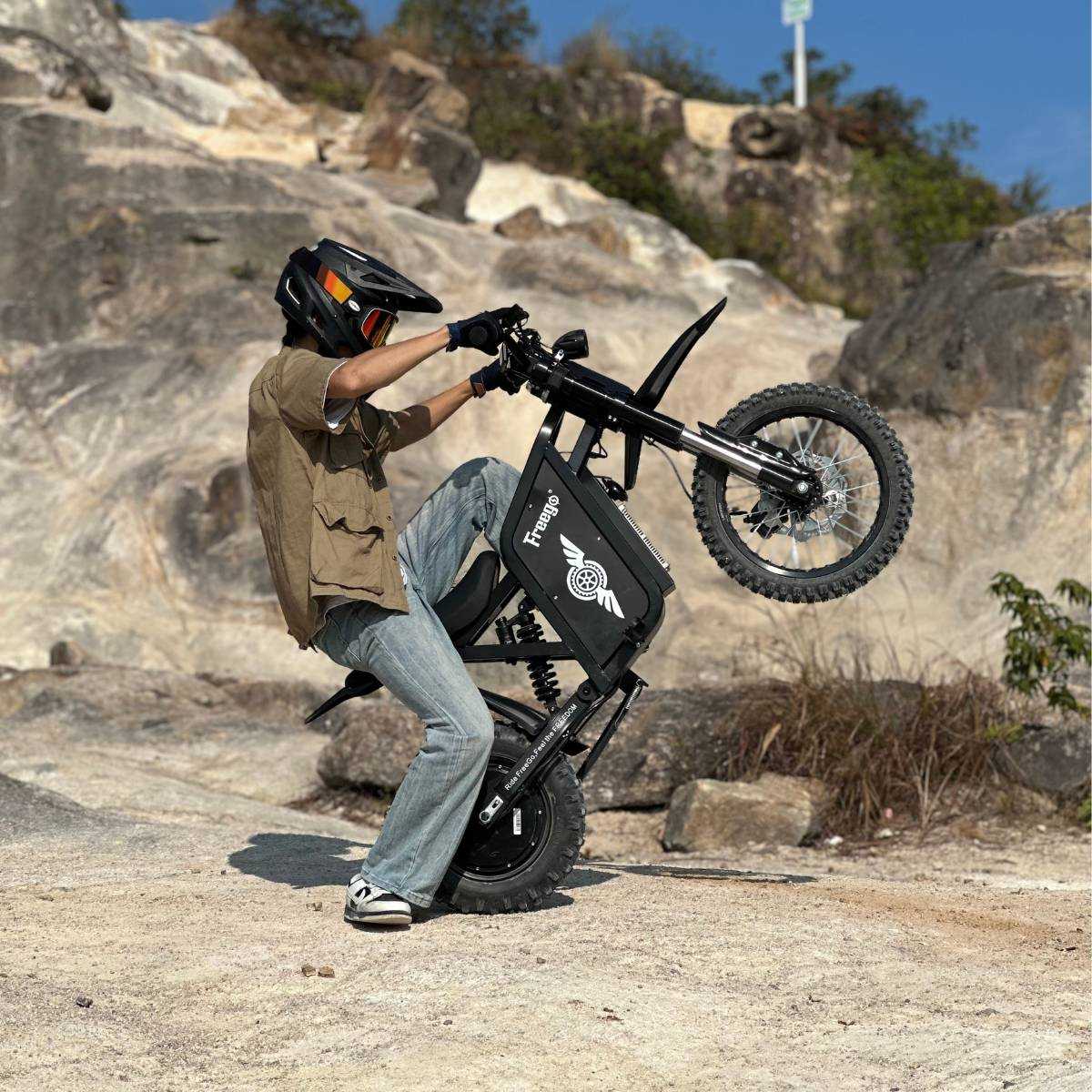 Helmeted rider doing a wheelie on a Freego-branded X0 electric mini dirt bike, set on sunlit rocky terrain.